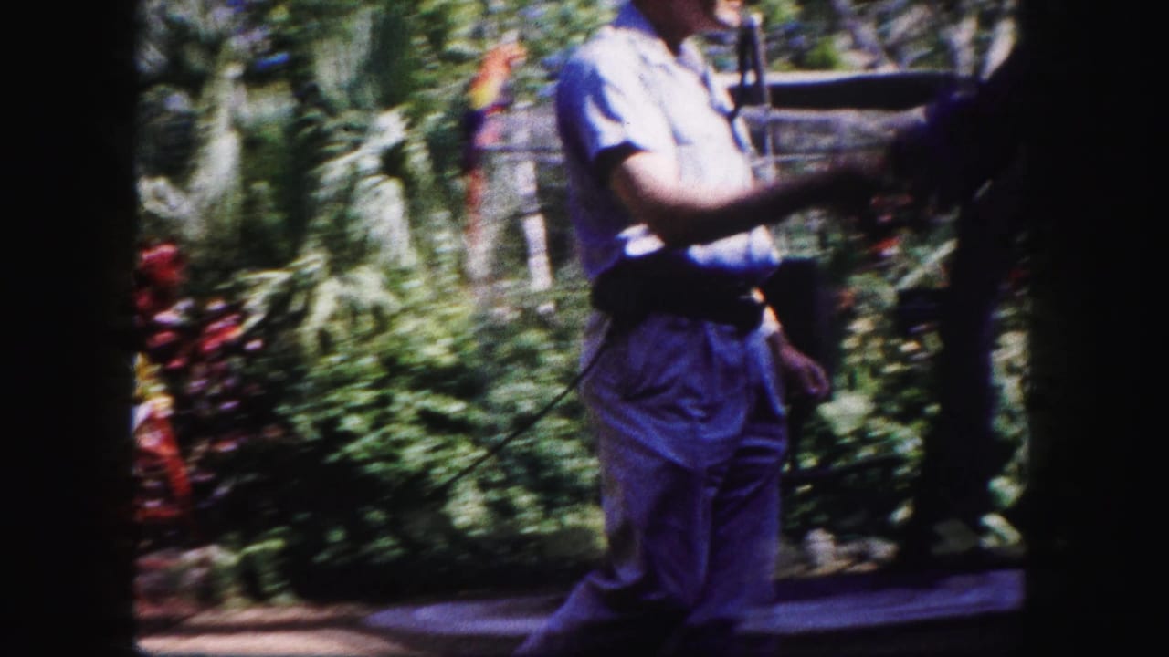 Man walks through a leafy zoo area carrying a long pole with a parrot enclosure nearby.