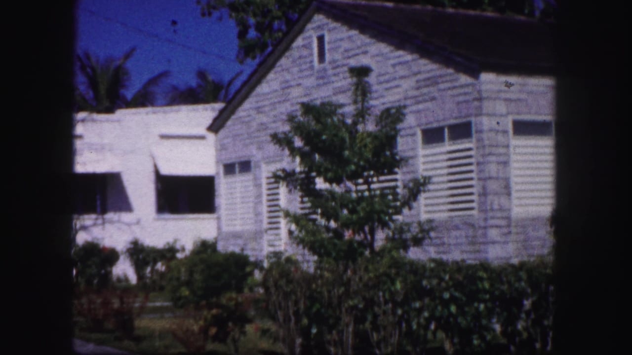 1960 Tampa home-movie frame showing white residential buildings, louvered shutters, shrubs, and palm foliage under a blue sky.