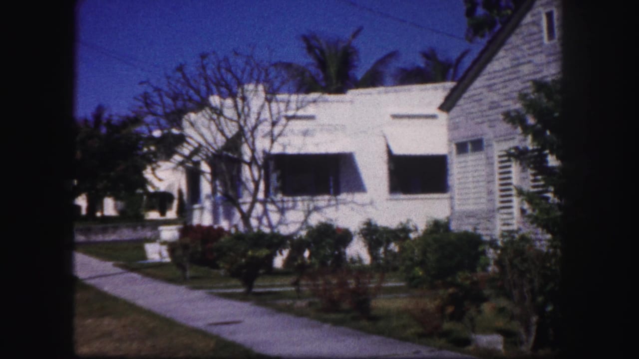 1960 Tampa street-side view with sidewalk, white house, dark awnings, leafless tree, shrubs, and part of a neighboring structure.