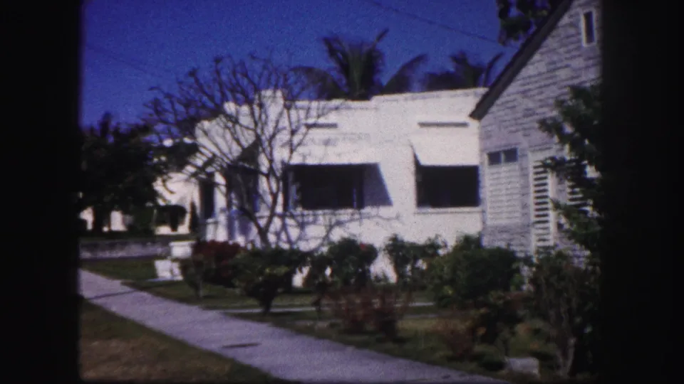 1960 Tampa street-side view with sidewalk, white house, dark awnings, leafless tree, shrubs, and part of a neighboring structure.