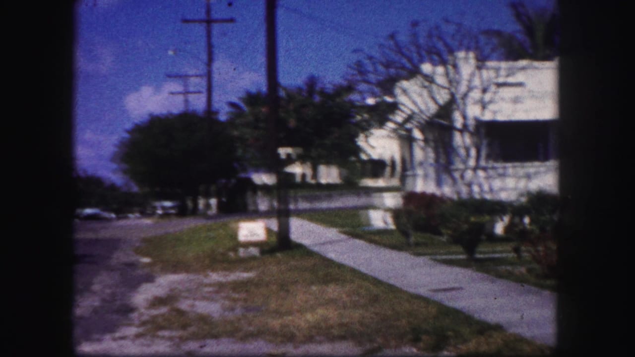 1960 Tampa home-movie frame showing a residential sidewalk, street edge, utility poles, lawn, trees, and a white house with awnings.