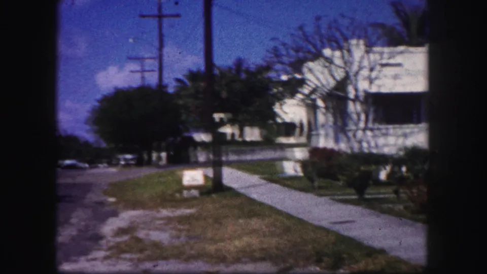1960 Tampa home-movie frame showing a residential sidewalk, street edge, utility poles, lawn, trees, and a white house with awnings.