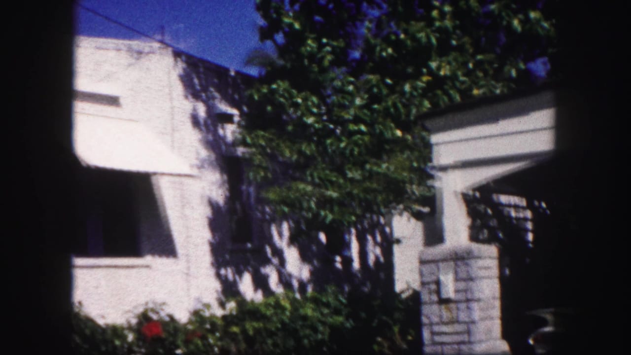 White stucco house exterior with shaded windows, shrubs, tree foliage, and a low wall near the entrance.