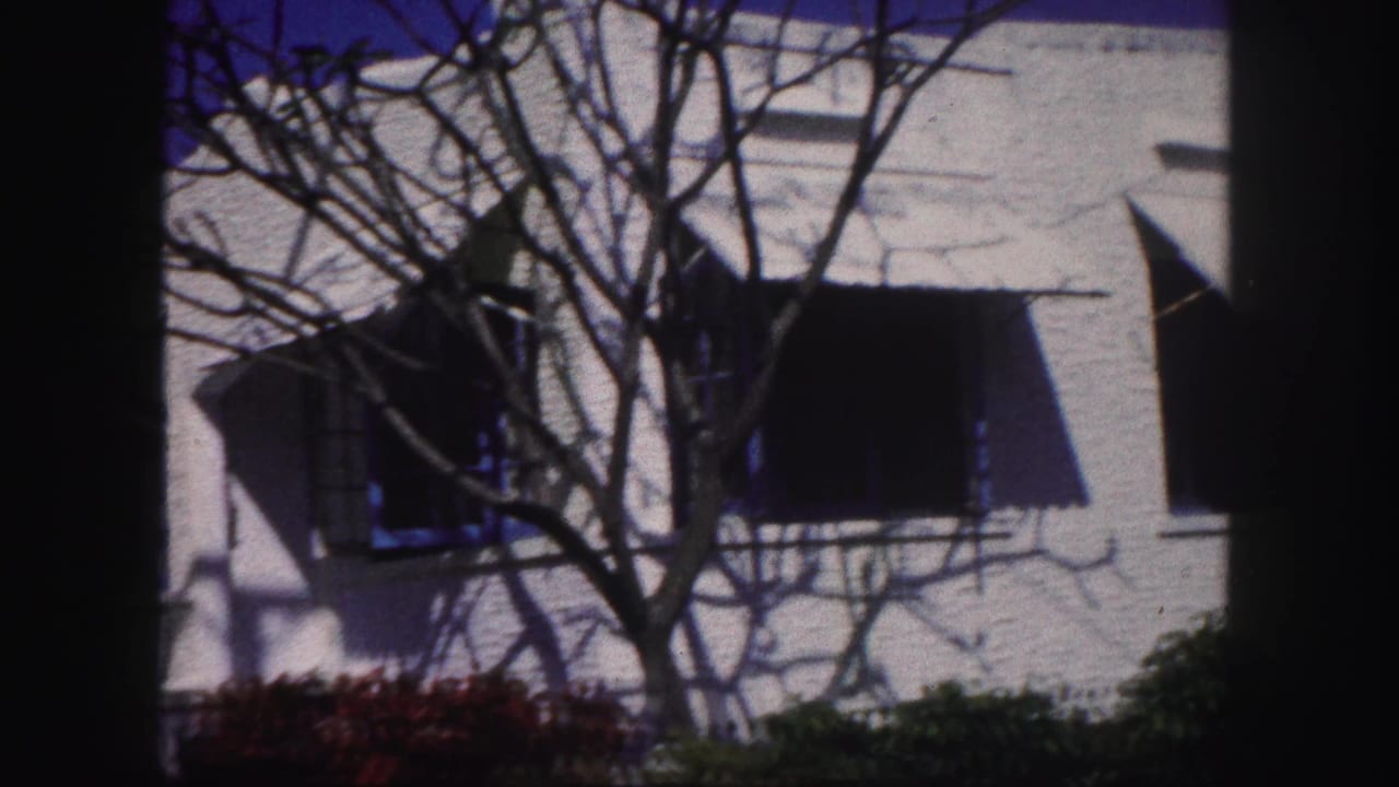 1960 Tampa home-movie frame showing a white stucco residential exterior with dark awnings, leafless branches, and strong sun shadows.