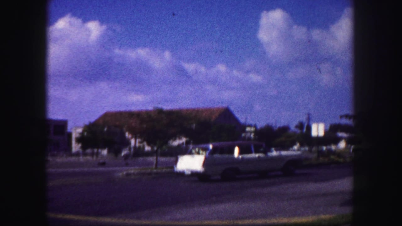 1960 Tampa street frame with a light-colored convertible passing a low building beneath a saturated blue sky and clouds.