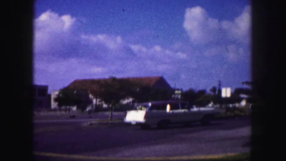 1960 Tampa street frame with a light-colored convertible passing a low building beneath a saturated blue sky and clouds.