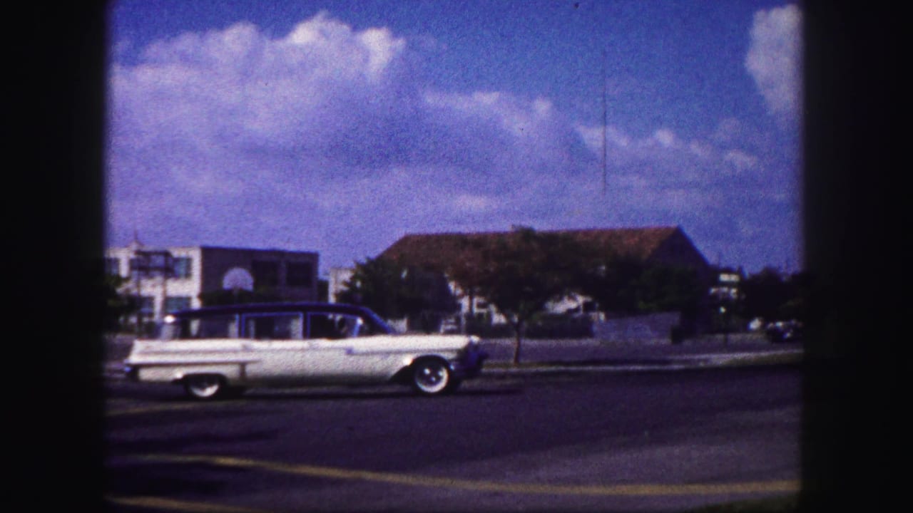 1960 Tampa home-movie frame of a white station wagon moving past low buildings, trees, pavement, and blue sky.