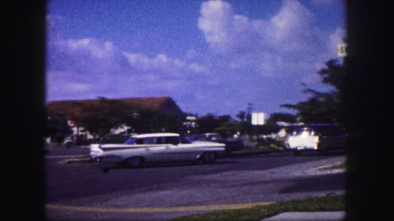 1960 Tampa home-movie frame of a white station wagon on a broad roadway with trees, lawns, and low buildings nearby.
