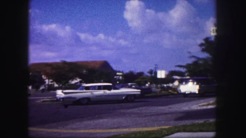 1960 Tampa home-movie frame of a white station wagon on a broad roadway with trees, lawns, and low buildings nearby.