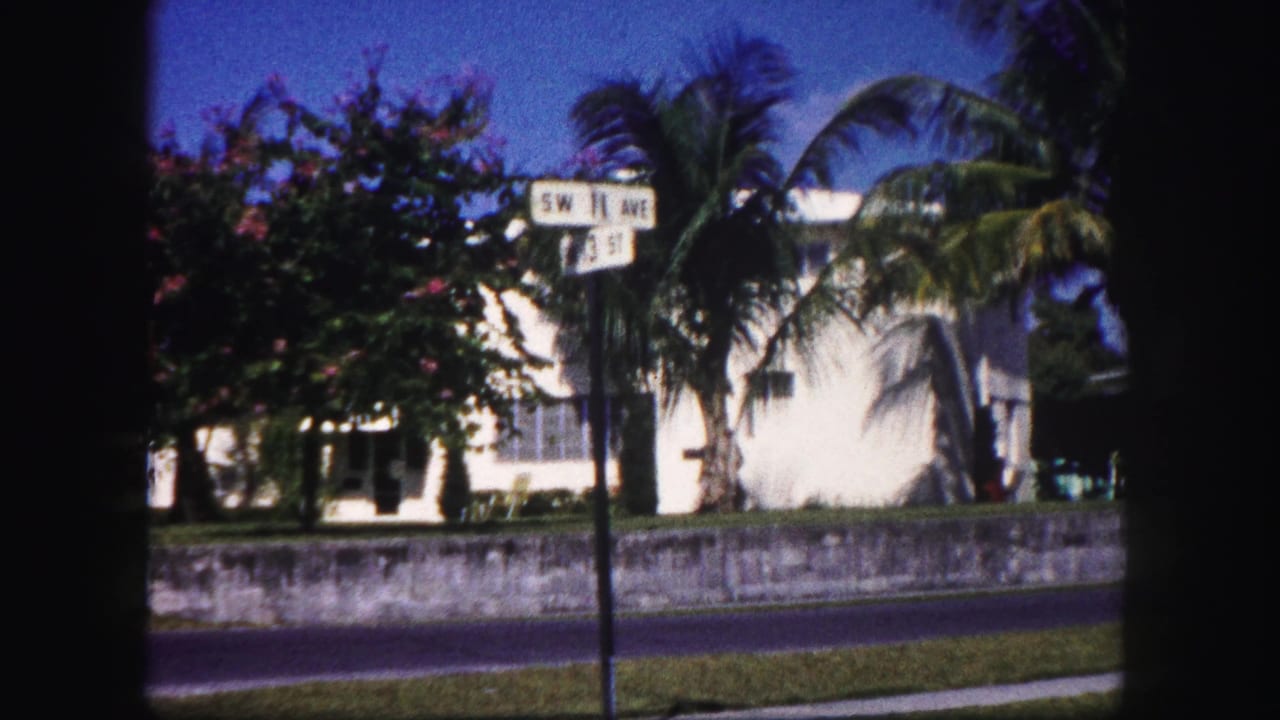 1960 Tampa home-movie frame showing the S 8th Ave and 2nd St sign, palm trees, a low wall, and white houses.