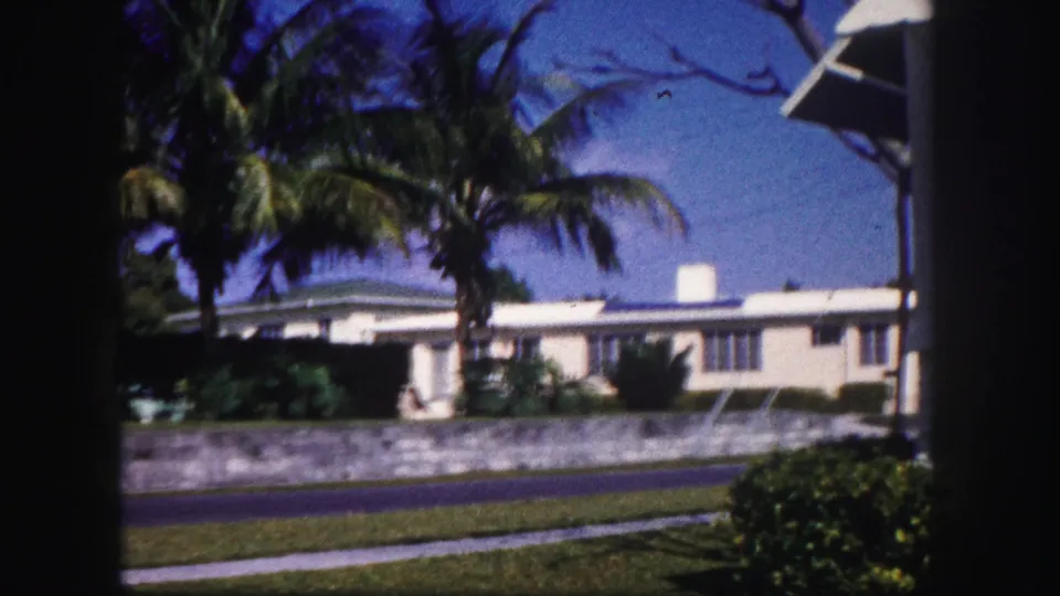1960 Tampa frame with a low white residence, palm trees, roadside grass, sidewalk, and dark film borders.