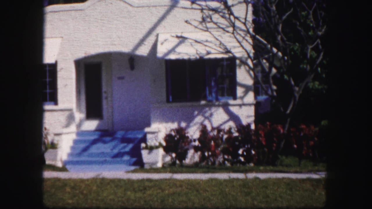 1960 Tampa home-movie view of a white stucco house with blue steps, dark shutters, shrubs, lawn, and tree shadows.