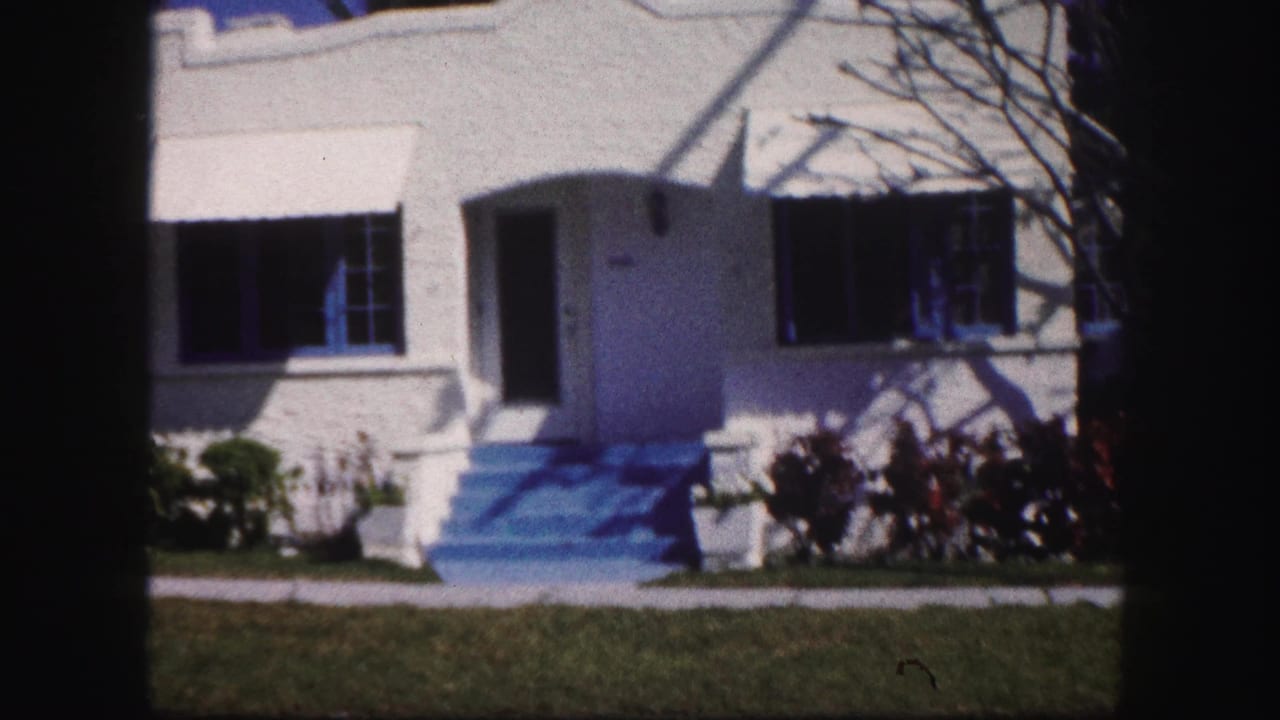 1960 Tampa frame showing a white stucco residence with blue-painted steps, dark shutters, front lawn, and garden shrubs.