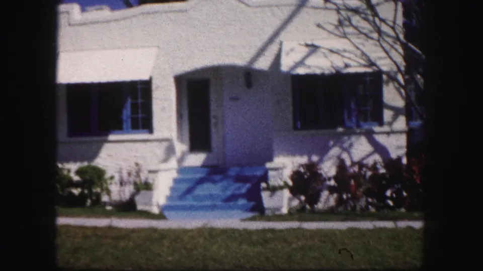 1960 Tampa frame showing a white stucco residence with blue-painted steps, dark shutters, front lawn, and garden shrubs.