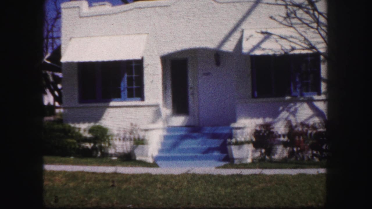 1960 Tampa home-movie frame showing a white stucco house with blue-painted steps, dark awnings, lawn, shrubs, and tree shadows.