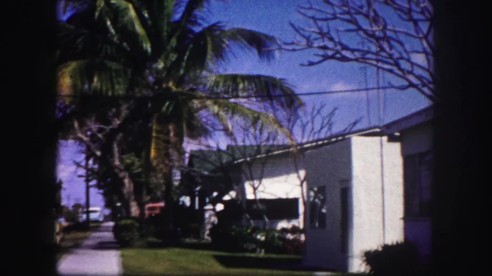 1960 Tampa home-movie frame showing a white stucco house, palm tree, awning, front lawn, and dark 8mm frame edge.