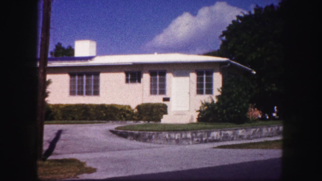 1960 Tampa home-movie view of a low white house with chimney, gridded windows, hedges, driveway, lawn, and bright sky.
