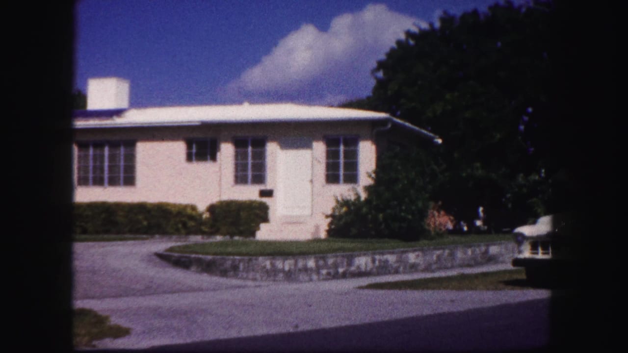 1960 Tampa residential frame showing a white one-story house, driveway, front hedges, large tree, and partial car at the right edge.