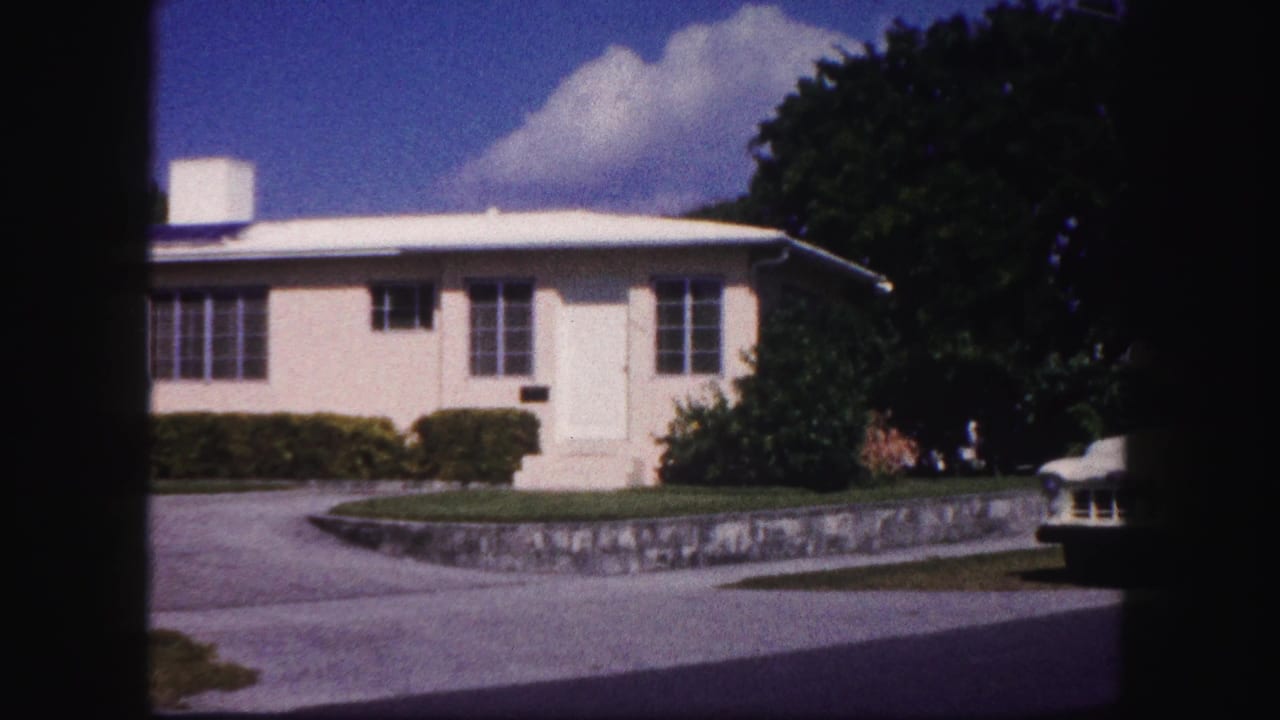 1960 Tampa home-movie frame showing a light stucco house beyond a curved driveway, with hedges, a stone retaining wall, trees, and blue sky.