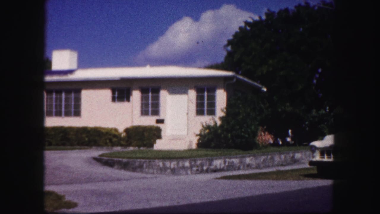 1960 Tampa frame with a pale house, low roofline, curved driveway, stone wall, dense trees, and the edge of a parked car visible at right.