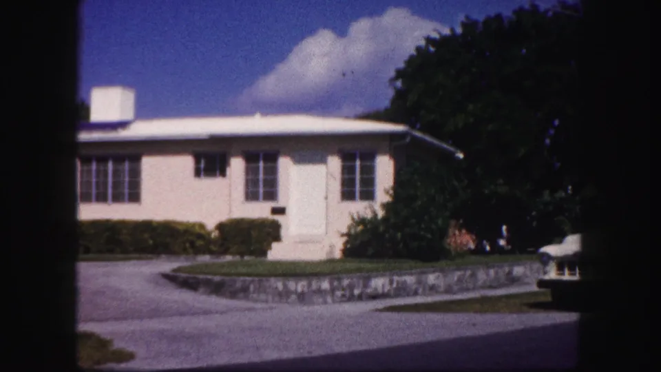 1960 Tampa frame with a pale house, low roofline, curved driveway, stone wall, dense trees, and the edge of a parked car visible at right.