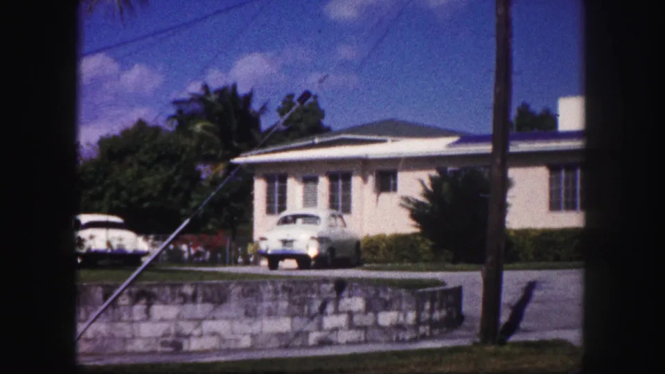 1960 Tampa home-movie view of the house frontage with light-colored cars, palm foliage, a utility pole, stone retaining wall, and driveway.