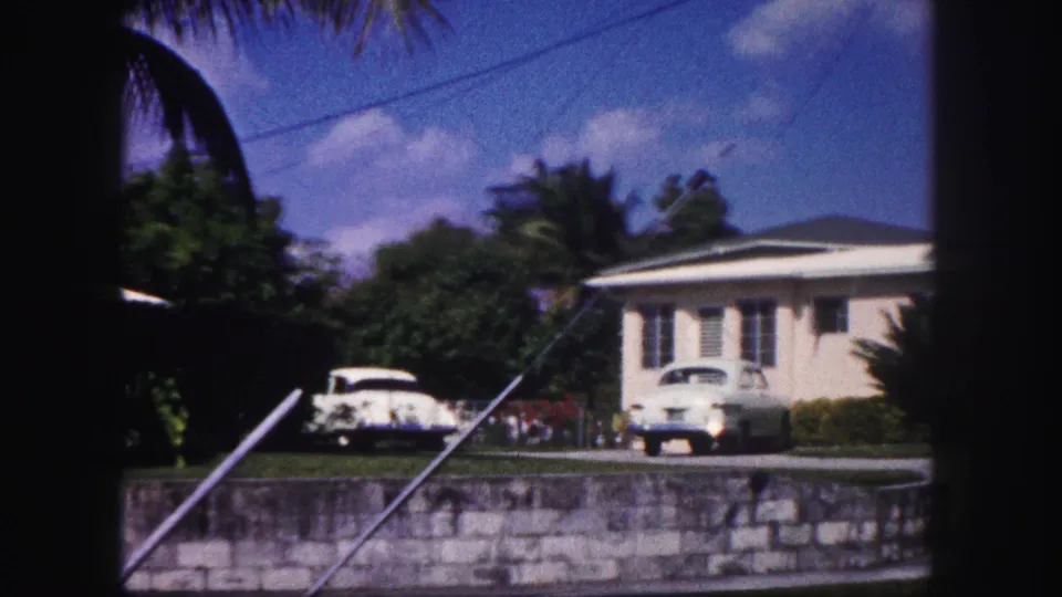 1960 Tampa frame showing a pale residential house with parked cars nearby, palm leaves overhead, utility lines, trees, driveway, and stone wall.