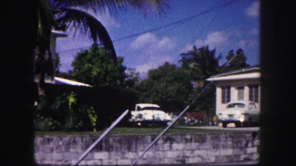 1960 Tampa home-movie frame showing a white suburban house, palms, lawn, low wall, and two parked mid-century cars.