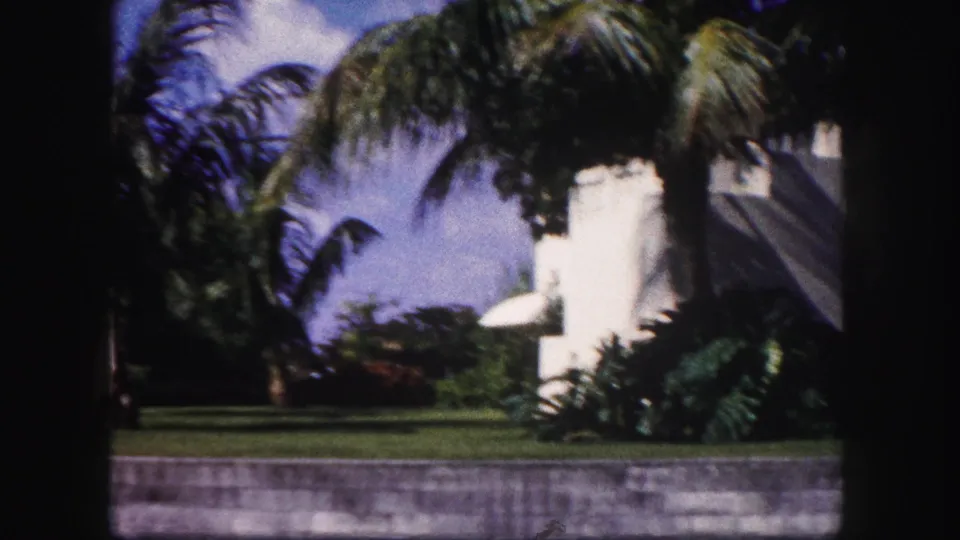 1960 Tampa home-movie view of palms, a white residential building, lawn, shrubs, blue sky, and the low wall along the street.