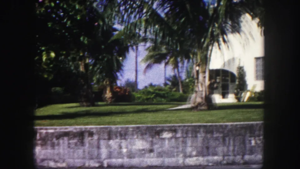 A white residence appears behind palms and shrubs, with a manicured lawn and low stone wall in 1960 Tampa home-movie footage.