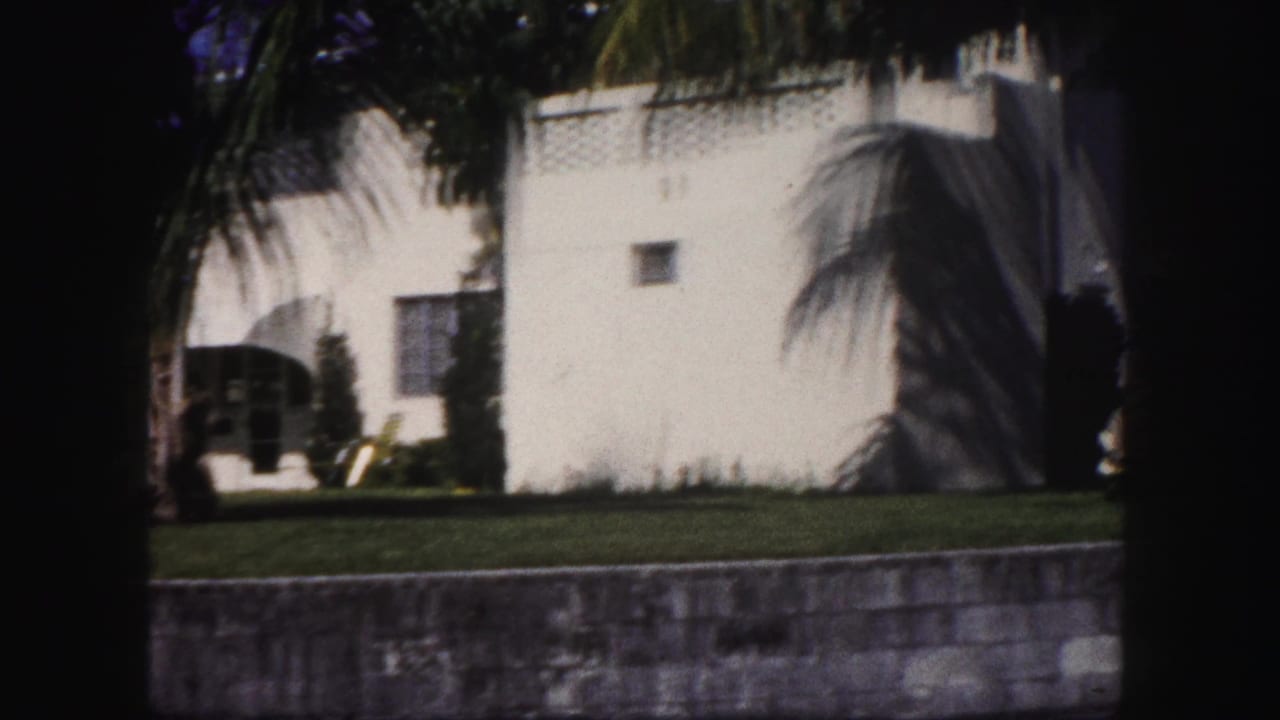 1960 Tampa home-movie frame showing a white stucco house, lawn, palm shadows, and a low wall in bright sun.