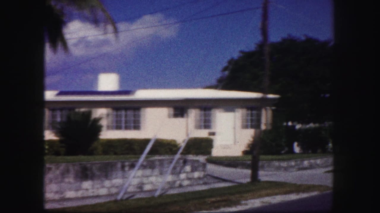1960 Tampa home-movie frame showing a white stucco house, blue trim, stone wall, lawn, shrubs, and bright residential street light.