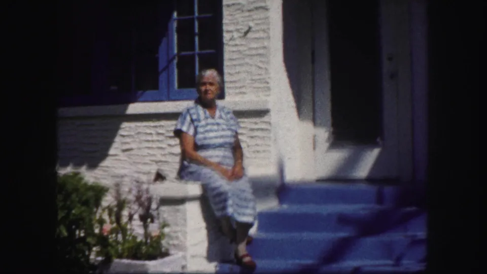 1960 Tampa home-movie frame of an older woman in a blue striped dress seated on front steps beside a stucco wall and blue-trimmed window.