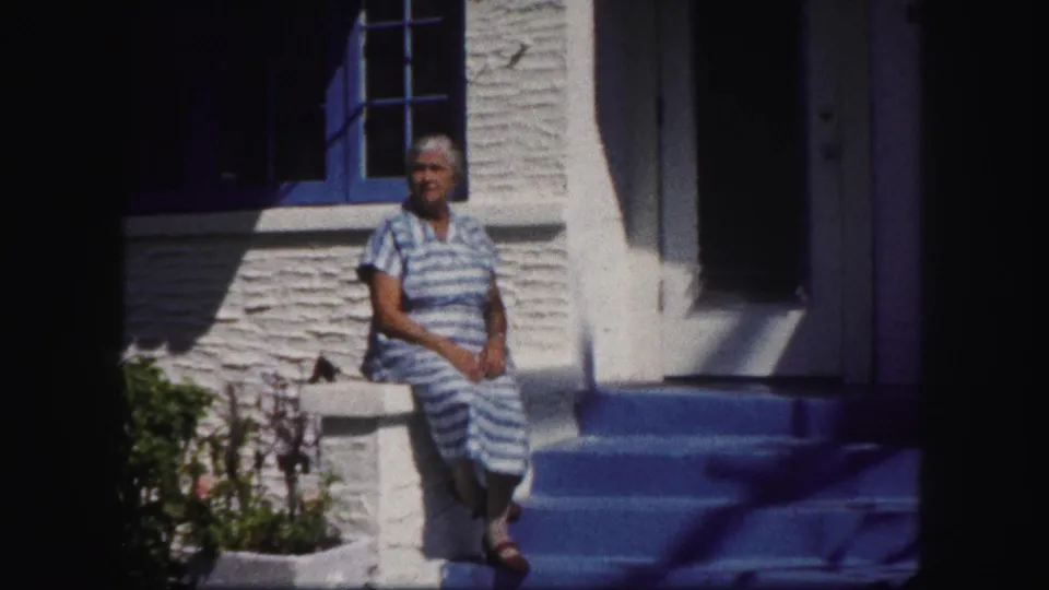 1960 Tampa home-movie frame showing an older woman seated outside a white stucco home, with blue trim, doorway, steps, and garden plants visible.