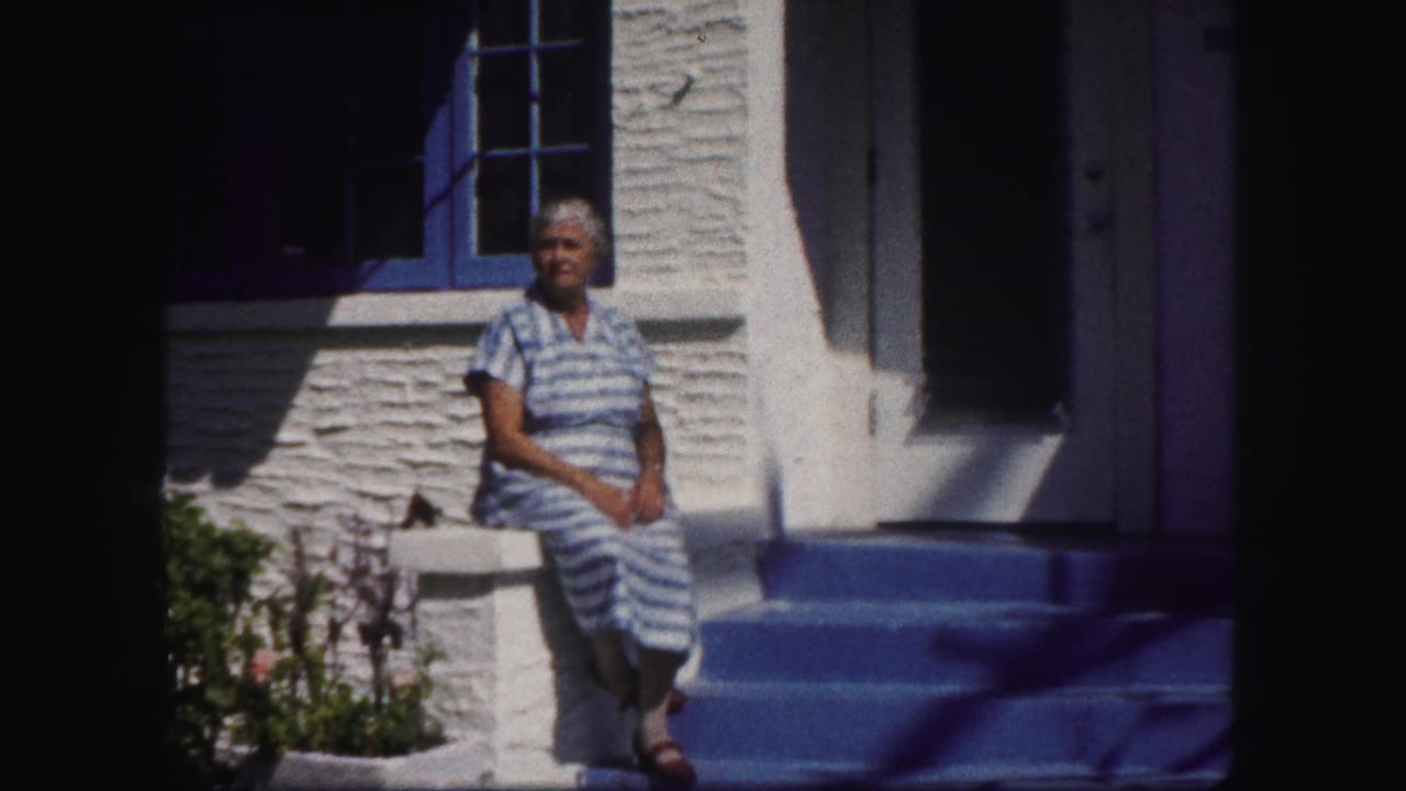In 1960 Tampa home-movie footage, a woman sits on porch steps beside blue-trimmed windows and a white stucco wall.