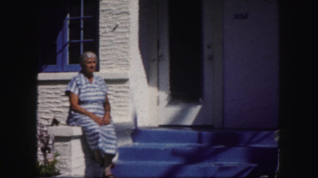 The woman remains seated outside the Tampa house, framed by a dark doorway, blue shutters, and bright stucco in harsh sun.