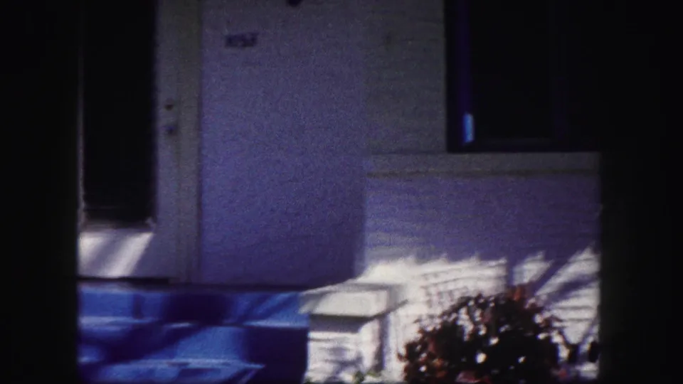 The camera view shifts away from the woman to the white stucco porch, blue trim, dark entrance, steps, and front garden.