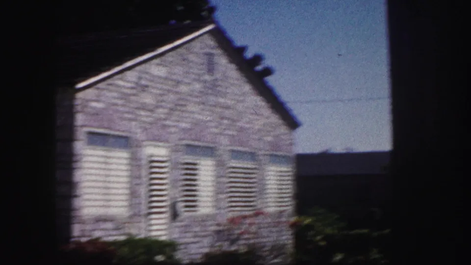 1960 Tampa side exterior view of a pale house with shutters, roofline, shrubs, and open blue sky.