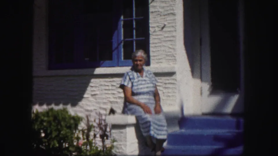 In 1960 Tampa home-movie footage, a woman in a blue striped dress sits on a white railing beside a blue-trimmed window.