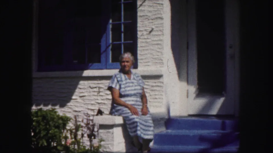 The woman remains seated outside the Tampa house, framed by white stucco, blue trim, and a dark doorway in strong sunlight.