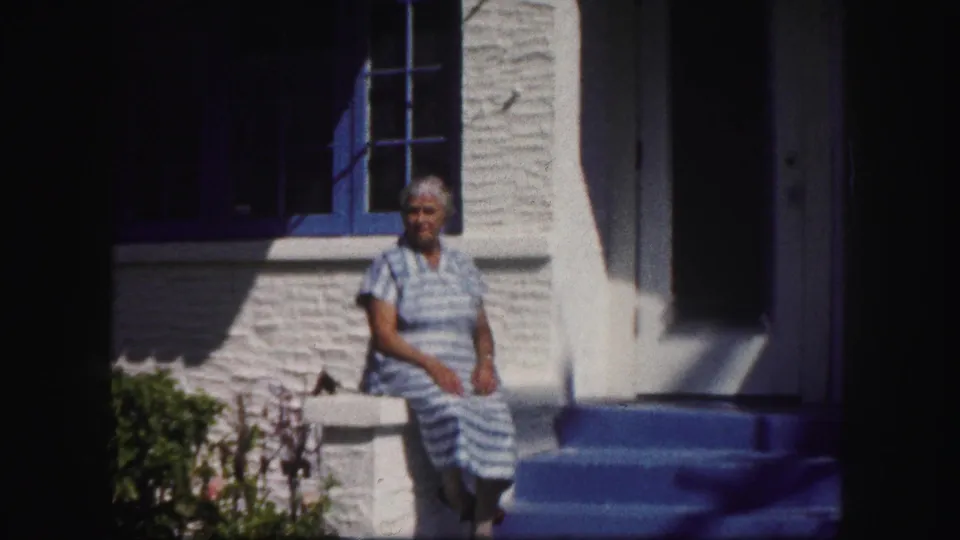 A home-movie frame shows the woman seated on the white railing, with blue steps, a window, and garden plants around the entry.