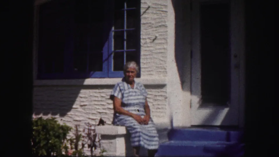 The final frame holds on the woman seated outside the white stucco house, with blue trim and the shaded doorway behind her.