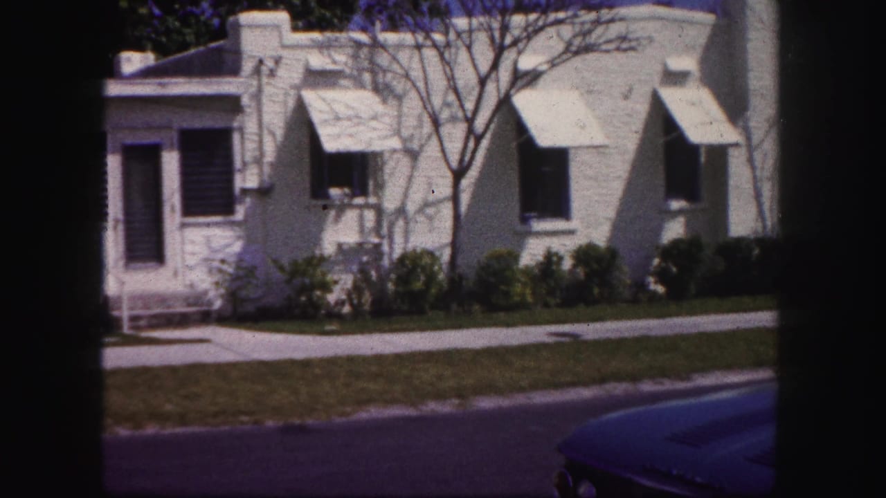 1960 Tampa home-movie frame showing a white stucco house with awnings, a front lawn, sidewalk, shrubs, and part of a blue car.