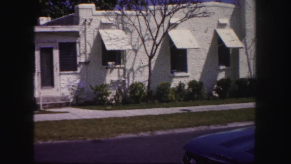 1960 Tampa home-movie frame showing a white stucco house with awnings, a front lawn, sidewalk, shrubs, and part of a blue car.