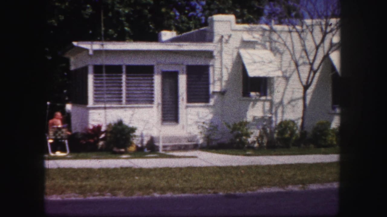 Front of a white house with louvered windows, awnings, shrubs, lawn, and a seated person near the entry.