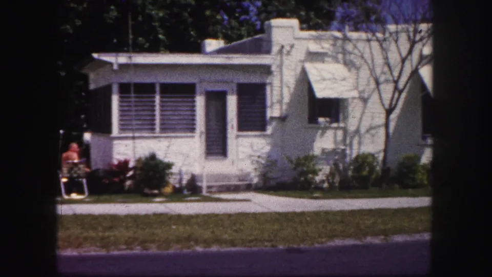 1960 Tampa frame showing a white stucco residence with louvered windows, awnings, lawn, shrubs, and a person seated outside.