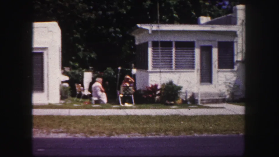 1960 Tampa home-movie frame with two adults seated in the yard near a white stucco house with louvered windows and shrubs.