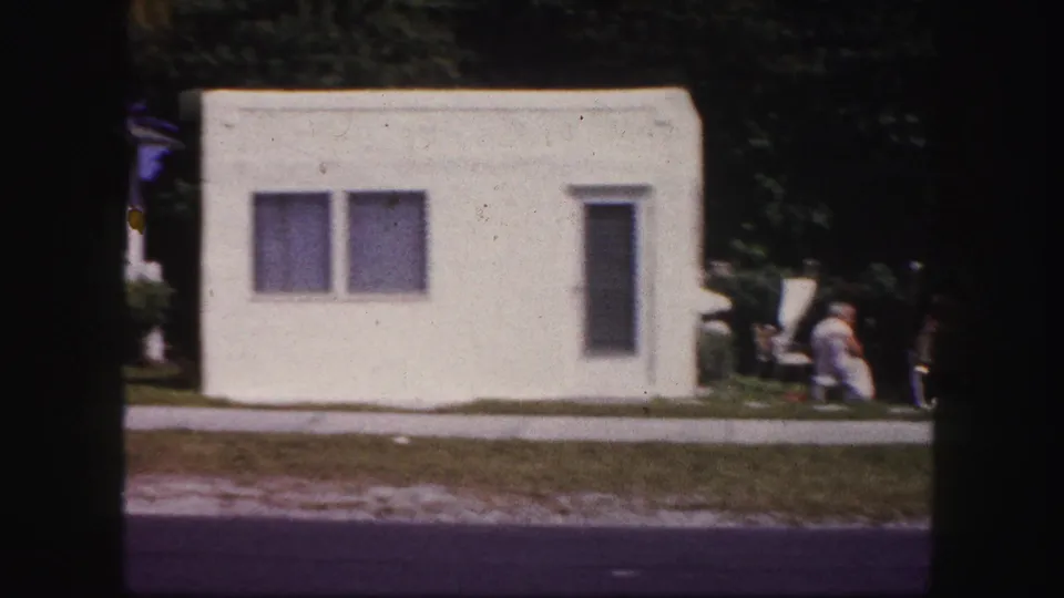 1960 Tampa frame showing the side of a white stucco house with two windows, lawn, sidewalk, and adults partly visible at the edge.