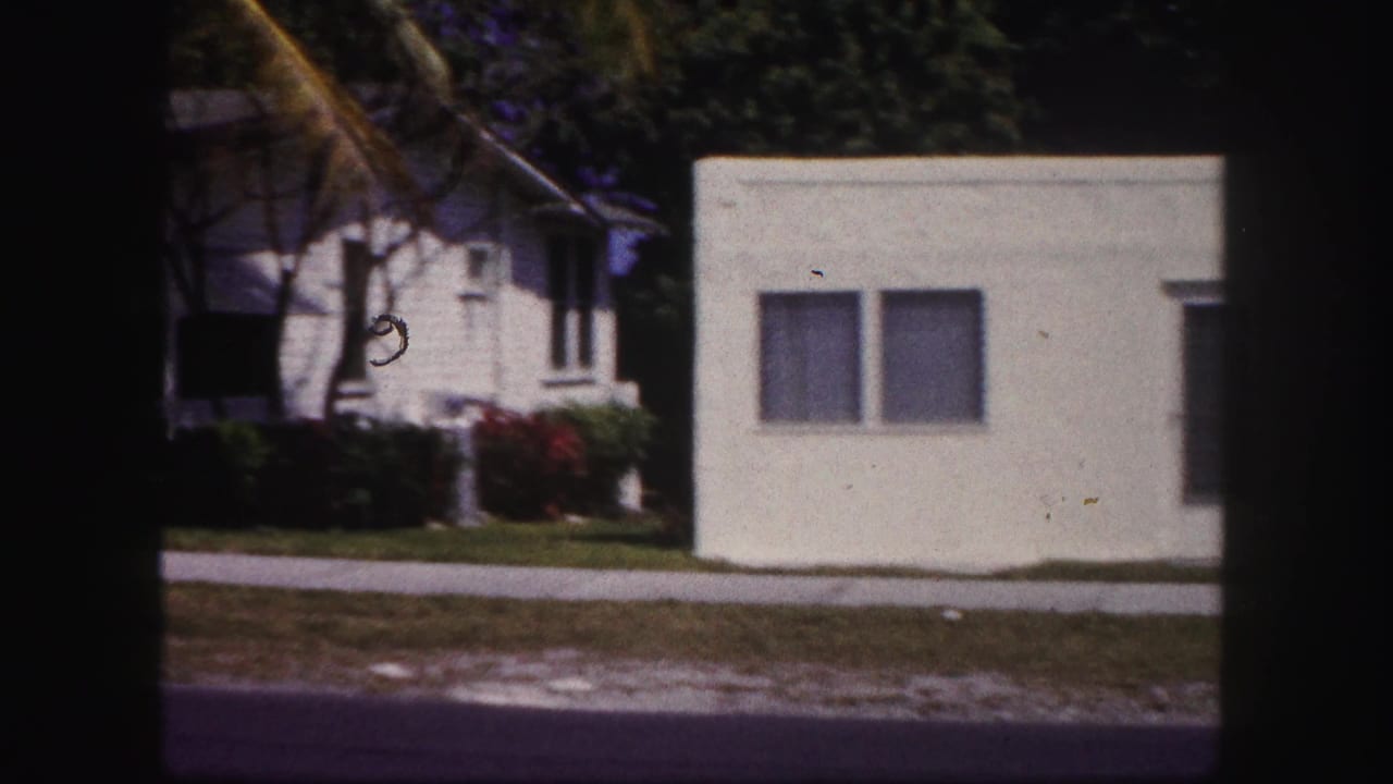 1960 Tampa home-movie frame showing white stucco houses, shaded windows, lawn, sidewalk, and roadside foreground.