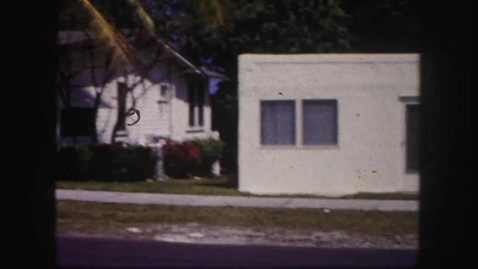 1960 Tampa home-movie frame showing white stucco houses, shaded windows, lawn, sidewalk, and roadside foreground.